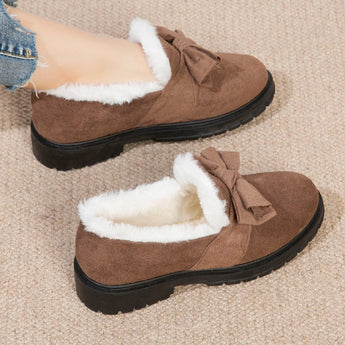 Women's faux fur-lined suede loafers in brown, featuring a bow tie design and moccasin style on a beige and grey wooden background.