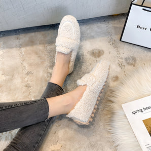 Beige women's fluffy slippers with a plush, non-slip sole on a wooden floor, designed for warmth and comfort during winter.