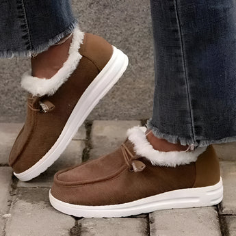 Women's brown fleece-lined winter loafers on a person's feet, styled with blue jeans; the loafers are warm slip-ons suitable for cold weather.
