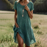 A woman wearing a green polka dot short summer dress made of lightweight bohemian chiffon, standing with one hand on her hip. The dress features short sleeves and a V-neckline, fitting comfortably around the waist. She is accessorized with sunglasses and surrounded by plants. The dress is in size 3XL.