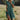 A woman wearing a green polka dot short summer dress made of lightweight bohemian chiffon, standing with one hand on her hip. The dress features short sleeves and a V-neckline, fitting comfortably around the waist. She is accessorized with sunglasses and surrounded by plants. The dress is in size 3XL.