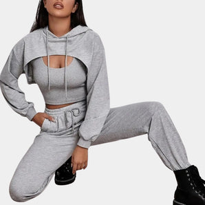 A woman modeling a grey tracksuit set featuring a cropped hoodie and joggers, styled for urban athleisure. She is posed in a seated position on a chair, showcasing the outfit's fit and comfort. The hoodie has long sleeves and a collar, while the joggers emphasize the waist and thighs.