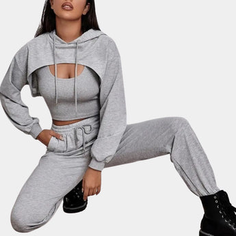 A woman modeling a grey tracksuit set featuring a cropped hoodie and joggers, styled for urban athleisure. She is posed in a seated position on a chair, showcasing the outfit's fit and comfort. The hoodie has long sleeves and a collar, while the joggers emphasize the waist and thighs.