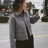 Woman wearing a stylish grey wool blend cropped button jacket, suitable for winter. The jacket features a fashionable design with a collar, long sleeves, and a fitted waist. The woman is standing indoors near a window, showcasing the jacket's elegant fit and design.