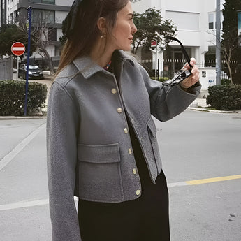 Woman wearing a stylish grey wool blend cropped button jacket, suitable for winter. The jacket features a fashionable design with a collar, long sleeves, and a fitted waist. The woman is standing indoors near a window, showcasing the jacket's elegant fit and design.