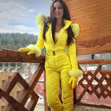 Woman wearing a stylish insulated yellow winter jumpsuit with a hood, designed for skiing, standing outdoors with a sky and clouds in the background.