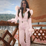 
A woman wearing a pink insulated hooded winter jumpsuit, smiling outdoors against a cloudy sky backdrop. The jumpsuit features a waist-cinching design, long sleeves, and a cozy collar, making it an ideal stylish choice for ski fashion.
