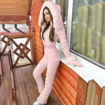Women's insulated black hooded winter jumpsuit showcased on a mannequin, highlighting its stylish ski fashion design. The jumpsuit features a waist-cinching silhouette and is made from textile material, perfect for leisure activities and fashionable winter wear. The image captures the jumpsuit standing against a wooden backdrop, with flash photography emphasizing its beauty and fashion-forward design.