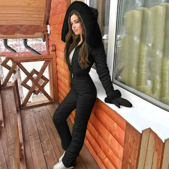 Women's insulated black hooded winter jumpsuit in size 5XL, showcasing a stylish ski fashion design. The outfit features a sleek black color, ideal for winter and outdoor activities. The image highlights the jumpsuit's outerwear quality, complemented by a modern hairstyle, set against a wooden backdrop.