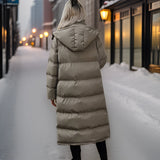 
A taupe-colored women's hooded puffer coat with a long design, featuring insulated fabric for warmth in winter conditions. The coat is styled with a waist-length fit and fur trim on the hood, suitable for freezing temperatures and snow. The sleeves are long, and the coat resembles a trench coat style, covering the legs for added protection against the cold.