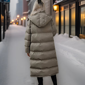 
A taupe-colored women's hooded puffer coat with a long design, featuring insulated fabric for warmth in winter conditions. The coat is styled with a waist-length fit and fur trim on the hood, suitable for freezing temperatures and snow. The sleeves are long, and the coat resembles a trench coat style, covering the legs for added protection against the cold.