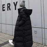 A woman wearing a long black puffer coat with a hood, sitting on a doorstep. The coat is an urban winter jacket featuring long sleeves and a fur-trimmed hood. The background includes a door, giving a street-style vibe to the setting.
