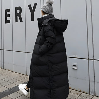 A woman wearing a long black puffer coat with a hood, sitting on a doorstep. The coat is an urban winter jacket featuring long sleeves and a fur-trimmed hood. The background includes a door, giving a street-style vibe to the setting.