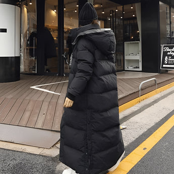 A woman wearing a long black puffer coat with a hood, walking on a city sidewalk.