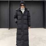 
A woman wearing a long black puffer coat with a hood, suitable for urban winter wear. The coat features a high collar and full sleeves. The woman is accessorized with sunglasses, emphasizing the coat's stylish and contemporary design.