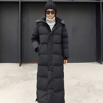 
A woman wearing a long black puffer coat with a hood, suitable for urban winter wear. The coat features a high collar and full sleeves. The woman is accessorized with sunglasses, emphasizing the coat's stylish and contemporary design.
