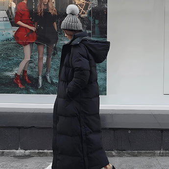 Women's long black puffer coat with hood, featuring a sleek urban design, worn by a woman standing and accessorized with a bag and eyewear.
