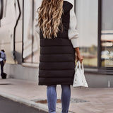 
A woman wearing a long black puffer vest styled as an urban chic winter jacket, featuring a sleek design with a waist-length fit. She is standing, showcasing the vest's shoulder and sleeve details.