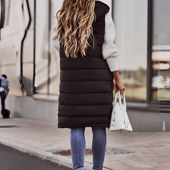 
A woman wearing a long black puffer vest styled as an urban chic winter jacket, featuring a sleek design with a waist-length fit. She is standing, showcasing the vest's shoulder and sleeve details.