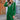 A woman wearing a long blue oversized zip-up hoodie stands outdoors. She is accessorized with sunglasses, and the hoodie features a large collar and long sleeves. The background is a blend of white and green elements, enhancing the urban streetwear vibe.