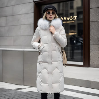 Women's long winter coat with a fur hood, featuring a quilted design, shown on a model. The coat is insulated for warmth and has a waist-cinching silhouette. The model is wearing sunglasses and a hat.