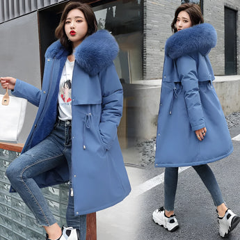 
A woman standing and modeling a long winter parka in blue, featuring a plush blue fur collar, adjustable waist, and long sleeves. The parka is designed as outerwear suitable for cold weather.