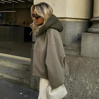
A woman wearing a luxurious oversized green high-collar hoodie, styled in an urban fashion. She pairs it with sleek sunglasses, embodying a modern and trendy look. The hoodie has long sleeves and extends to cover the waist and thighs, providing a comfortable and chic appearance.