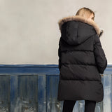 
A woman wearing a luxury white puffer jacket with a fur hood, standing outdoors. The jacket has long sleeves and is paired with denim jeans. The setting appears to involve wood, possibly a wooden deck or pathway. The image is taken with flash photography, highlighting the texture and details of the outerwear.