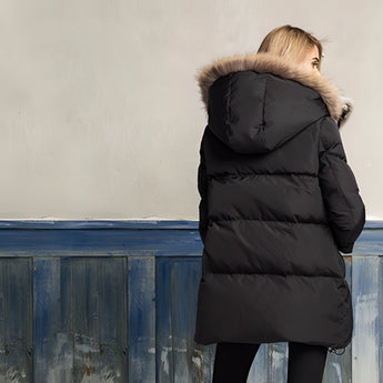 
A woman wearing a luxury white puffer jacket with a fur hood, standing outdoors. The jacket has long sleeves and is paired with denim jeans. The setting appears to involve wood, possibly a wooden deck or pathway. The image is taken with flash photography, highlighting the texture and details of the outerwear.