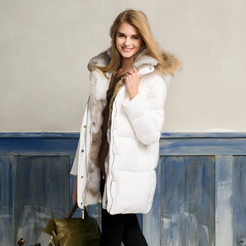 White puffer jacket with fur hood worn by a woman standing and smiling, showcasing the coat's luxurious design and winter style.