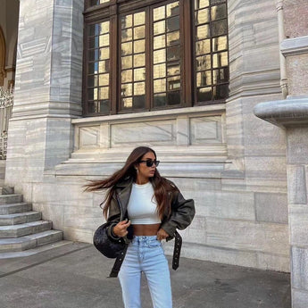 
A woman wearing an olive green belted biker leather jacket with an asymmetric zip, standing near a window. She is accessorized with sunglasses and a bag, highlighting the stylish and functional elements of the jacket.
