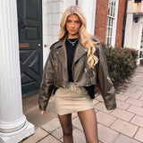 Women's olive green belted biker leather jacket with an asymmetric zip detail, featuring long sleeves and a stylish collar. The jacket is displayed on a mannequin or model against a neutral background, showcasing its waist belt and high-quality textile material.