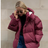 
A woman wearing a wine red oversized hooded puffer jacket with a trendy winter fashion design. She is smiling and appears happy, with the jacket featuring long sleeves and a waist length.