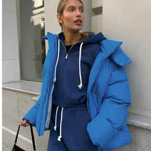 
A woman wearing an oversized blue hooded puffer jacket, showcasing trendy winter fashion. The jacket features a bright electric blue color with long sleeves and an ample fit, highlighting the shoulder and waist areas. The woman is also carrying a stylish bag, complementing her fashionable ensemble.
