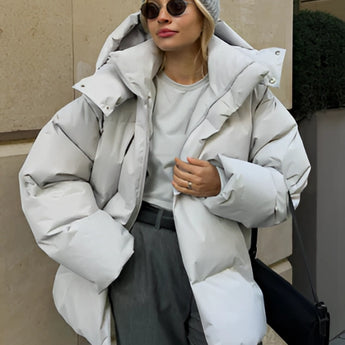
A model wearing a women's oversized blue hooded puffer jacket, featuring long sleeves and a drawstring hood, styled for trendy winter fashion. The jacket complements a pair of stylish sunglasses, highlighting the fashion-forward look. The jacket's large fit provides a comfortable and warm choice for cold weather attire.