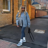 A woman wearing an oversized gray wool blend coat with chunky knit detailing, styled for winter fashion. She is dressed in jeans and shoes, accessorized with eyewear and a bag. The scene includes a window and road surface in the background.
