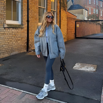 A woman wearing an oversized gray wool blend coat with chunky knit detailing, styled for winter fashion. She is dressed in jeans and shoes, accessorized with eyewear and a bag. The scene includes a window and road surface in the background.