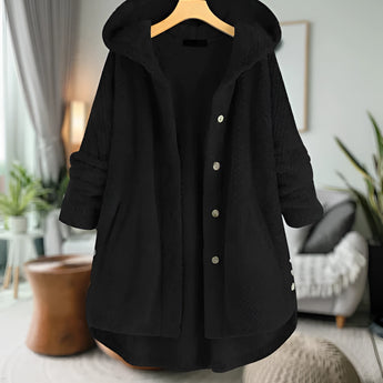 Women's oversized wool hooded sweater coat in Nero color, displayed in a cozy winter setting. The coat features a loose fit with long sleeves and a high collar, providing warmth and comfort. A potted houseplant adds a touch of greenery to the scene. The sweater coat is designed for plus-size fashion, available in 5XL.