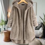 Woman wearing a beige oversized wool hooded sweater coat, displayed on a clothes hanger, with a cozy winter design. The coat is hanging in a room decorated with potted plants and houseplants in the background. The sweater features a collar and long sleeves.