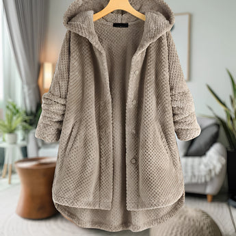 Woman wearing a beige oversized wool hooded sweater coat, displayed on a clothes hanger, with a cozy winter design. The coat is hanging in a room decorated with potted plants and houseplants in the background. The sweater features a collar and long sleeves.