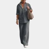 A woman wearing a gray linen suit consisting of a casual button-down shirt and matching pants, suitable for summer. The shirt features a collar and includes long sleeves, while the fit is relaxed, ideal for a casual setting. The woman may be holding a bag, enhancing the casual and functional style of the outfit.
