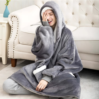 
A woman sitting comfortably on a couch wearing a gray oversized fleece hooded winter robe, showcasing its plush texture and warmth. The robe drapes over her knees and thighs, highlighting its relaxed fit and cozy design for lounging.