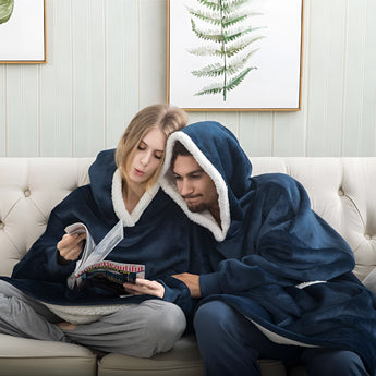 
A woman comfortably sitting on a couch in a living room, wearing a dark blue plush oversized fleece hooded winter robe, exuding warmth and coziness, with a picture frame visible in the background.