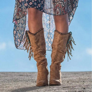 Tan suede mid-calf boots with fringe detail, worn by a person standing outdoors, partially visible with a dress and surrounded by natural scenery.