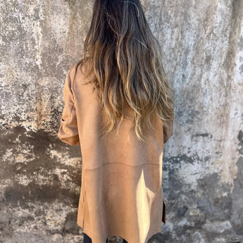 A woman wearing a tan suede blazer with a lightweight design, perfect for fall fashion, standing outdoors in sunlight. The jacket features a tailored fit with long sleeves and is styled over a casual grey T-shirt. The woman's hair and shoulders are visible, complementing the chic and versatile look.