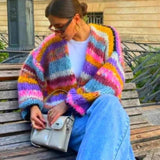 Woman wearing oversized multicolor stripe knit cardigan in dark colors, with glasses and shoulder-length hair.