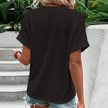 A woman wearing a white short sleeve V-neck blouse with a geometric pattern, paired with jeans, standing outdoors.