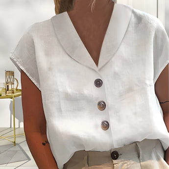 A women's white linen blouse with short sleeves, a V-neck, and button-down front, displayed on a chair.