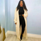 A woman wearing a cream-colored, oversized cable knit cardigan with long sleeves, styled as a cozy sweater. She has black hair and is posing in a relaxed manner. The cardigan falls to her thighs, emphasizing a casual and comfortable fashion design.