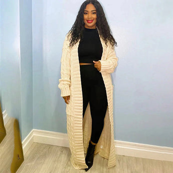 A woman wearing a cream-colored, oversized cable knit cardigan with long sleeves, styled as a cozy sweater. She has black hair and is posing in a relaxed manner. The cardigan falls to her thighs, emphasizing a casual and comfortable fashion design.